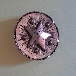 May include: A round metallic button with a copper-toned finish featuring a central five-pointed star. The surface is decorated with engraved radiating lines and four small circular studs positioned around the star. The edge has a textured rim.