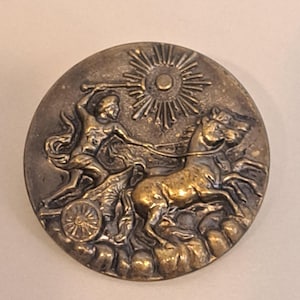 Peut inclure: Un bouton métallique rond présentant un relief représentant une silhouette conduisant un char tiré par des chevaux. Au-dessus du char, un soleil aux rayons rayonnants est illustré. Le bouton a une finition bronze antique patinée.