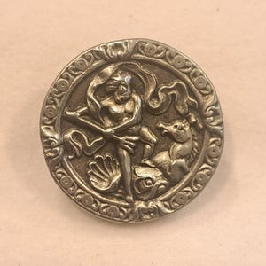 Puede incluir: Un botón circular de metal plateado con un diseño en relieve de una figura que sostiene un tridente, de pie junto a una criatura marina y una concha. El borde del botón está decorado con un patrón de volutas en relieve por todo el contorno.