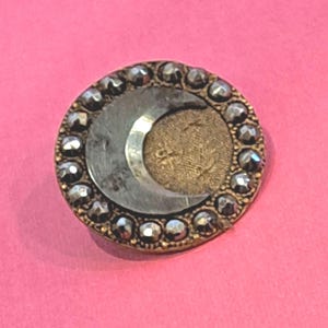 May include: A circular button featuring a central crescent moon shape in a metallic silver finish set against a textured gold-toned background. The outer edge is bordered by a ring of small, faceted, dark metallic studs arranged in a circular pattern.