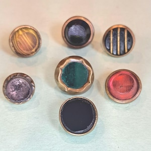 May include: A collection of seven circular buttons with metallic rims. The buttons feature various center designs including a yellow striped pattern, a black ribbed center, a black and gold striped design, a solid black disc, a red ribbed center, and a green center.