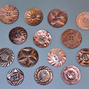 Puede incluir: Una colección de botones antiguos de color cobre con varios diseños en relieve. Los botones presentan patrones florales, geométricos y de estrella, cada uno con múltiples agujeros para coser. Los botones están dispuestos sobre una superficie azul claro.
