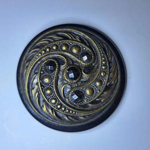 Op de afbeelding: Een ronde, sierlijke knoop met een zwarte basis en gouden accenten. De knoop heeft een wervelend ontwerp met ingebedde zwarte edelstenen en een gedetailleerd, gestructureerd patroon. Het ontwerp van de knoop is symmetrisch en opvallend.