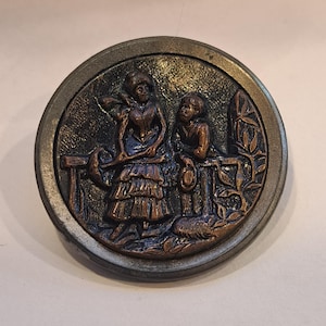 May include: A circular antique metal button featuring a detailed relief scene. The scene shows two figures, a woman and a child, near a fence. The button has a dark bronze centre and a silver-toned outer rim.