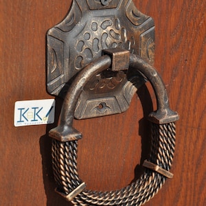 May include: A decorative door knocker with a rope-like design and a detailed, ornate base. The knocker is made of metal and has a dark brown finish.