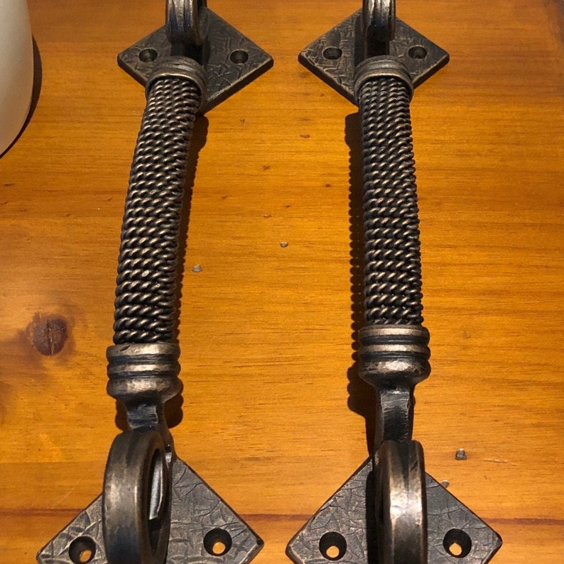 May include: Two antique-style door handles with a braided design. The handles are made of metal and have a dark brown finish. Each handle is attached to a square base with four holes.