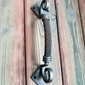 May include: A black metal door handle with a twisted rope design. The handle is attached to a square plate with a decorative swirl at the top and bottom.