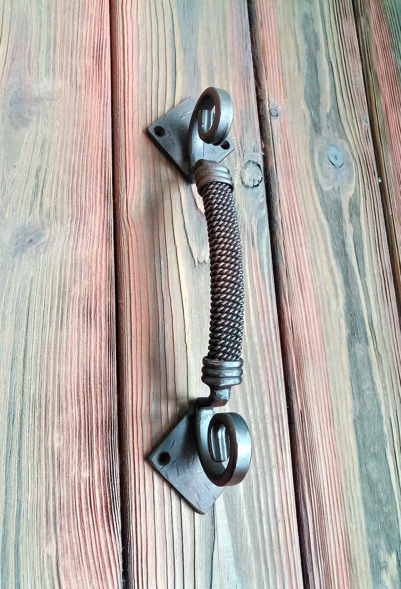 May include: A black metal door handle with a twisted rope design. The handle is attached to a square plate with a decorative swirl at the top and bottom.