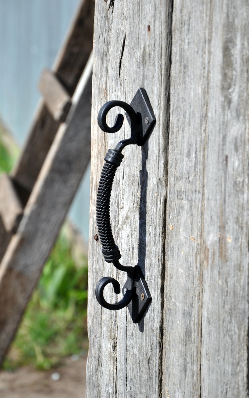 May include: Black metal door handle with a twisted rope design and decorative scrollwork. The handle is mounted on a weathered wooden door.