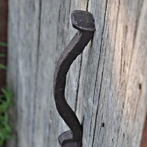 May include: A rustic, hand-forged iron door handle, shaped like a large nail, is mounted on a weathered wooden door.