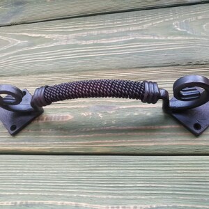 May include: Black metal door handle with a twisted rope design and decorative scroll ends. The handle is mounted on a square base.