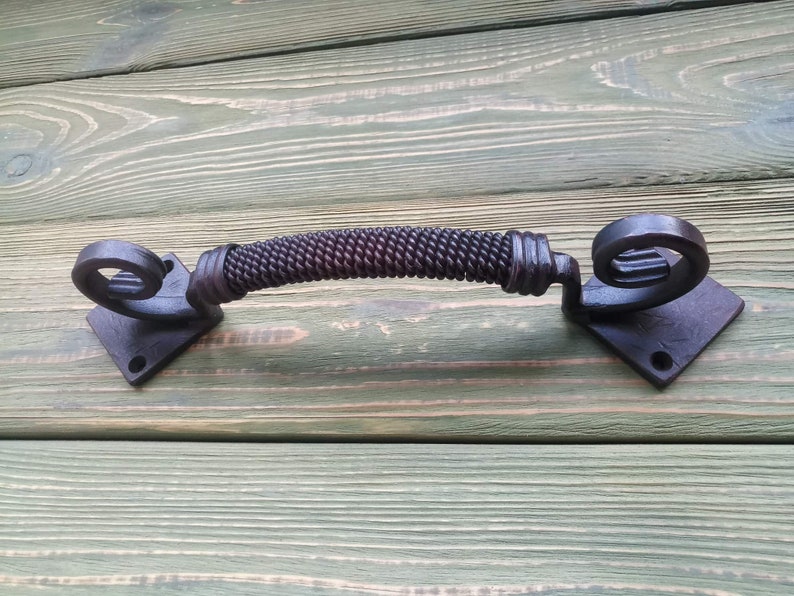 May include: Black metal door handle with a twisted rope design and decorative scroll ends. The handle is mounted on a square base.