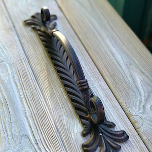 Könnte beinhalten: Ein schwarzer Türgriff aus Metall mit einem dekorativen, verzierten Design. Der Griff hat ein blattartiges Muster und eine Rolle oben.