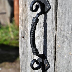 May include: Black metal door handle with a decorative, twisted design. The handle is attached to a wooden door.