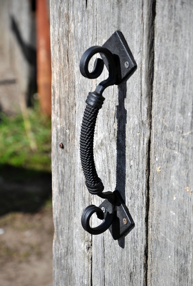 May include: Black metal door handle with a decorative, twisted design. The handle is attached to a wooden door.