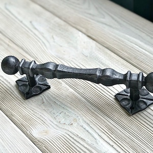 Peut inclure: Poignée de porte en métal noir avec des détails ornés et des boutons ronds. La poignée est montée sur une surface en bois blanc.