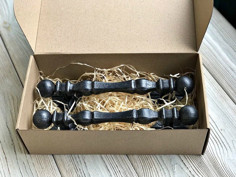 K&ouml;nnte beinhalten: Zwei schwarze Metall-Schubladengriffe mit abgerundeten Enden, verpackt in einem braunen Karton mit zerkleinertem Papier als F&uuml;llmaterial.
