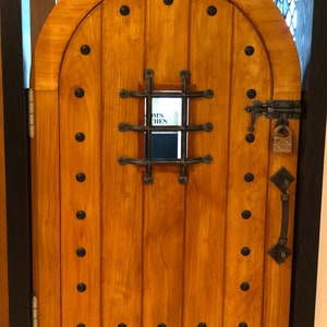 May include: A wooden arched door with a metal grate window and a metal handle. The door has a dark brown stain and is decorated with black metal rivets. The window has a white sign that reads "Amy's Kitchen".