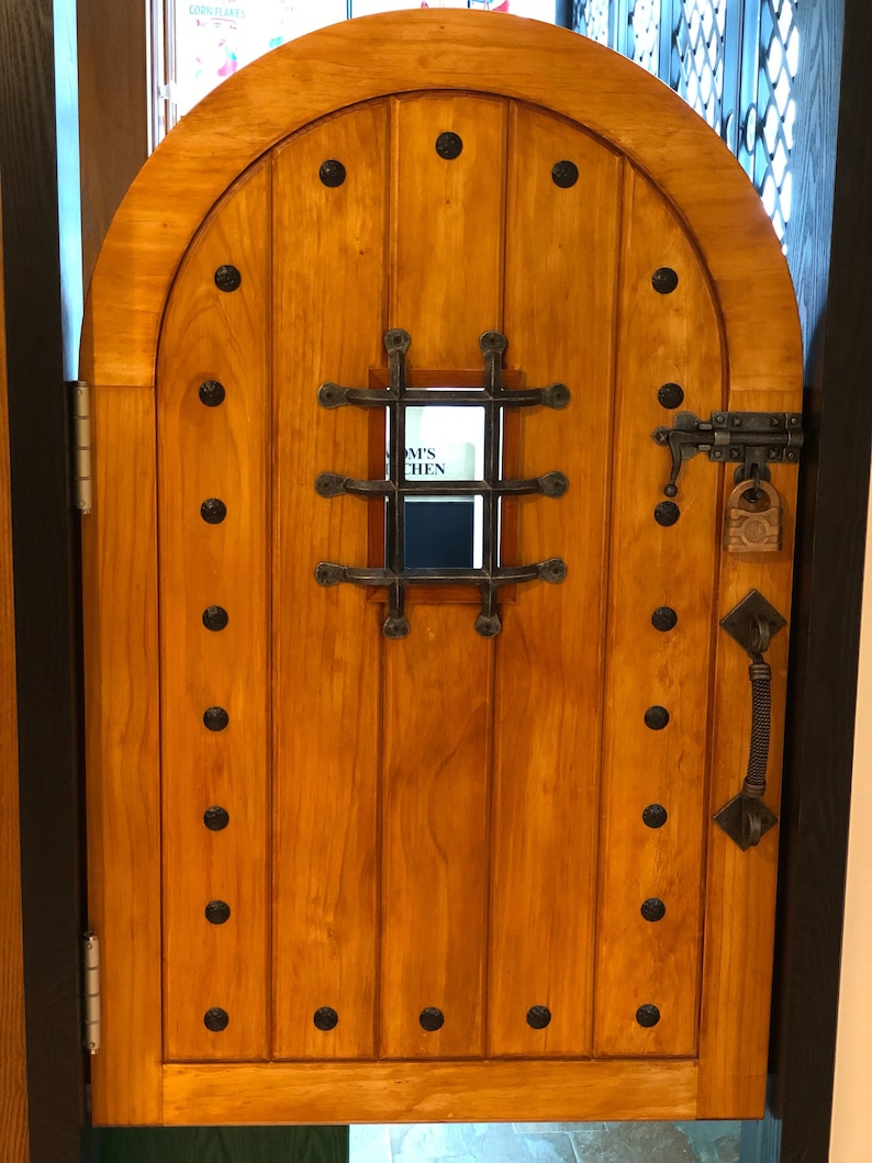 May include: A wooden arched door with a metal grate window and a metal handle. The door has a dark brown stain and is decorated with black metal rivets. The window has a white sign that reads "Amy's Kitchen".