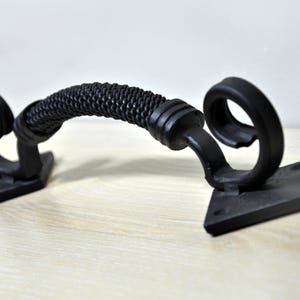 May include: Black metal door handle with a braided design on the handle and a scroll design on the ends. The handle is mounted on two square plates.