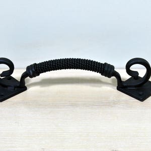 May include: Black metal door pull with a rope-like design and decorative scroll ends. The pull is mounted on square plates.