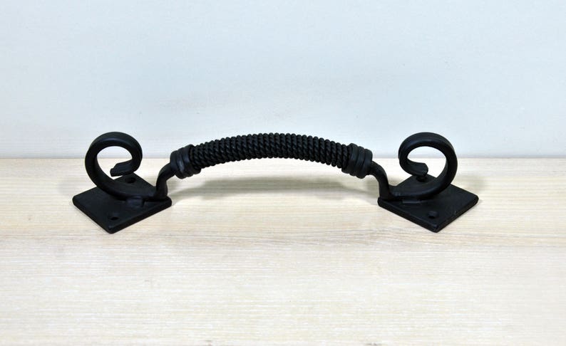 May include: Black metal door pull with a rope-like design and decorative scroll ends. The pull is mounted on square plates.