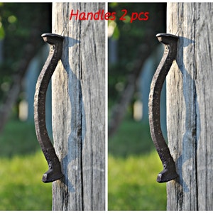 Può includere: Due maniglie in metallo rustico, ciascuna a forma di ferro di cavallo, sono fissate a un palo di legno invecchiato. Le maniglie sono di colore marrone scuro e hanno una superficie ruvida e testurizzata. Il testo "Handles 2 pcs" è stampato in rosso sopra le maniglie.