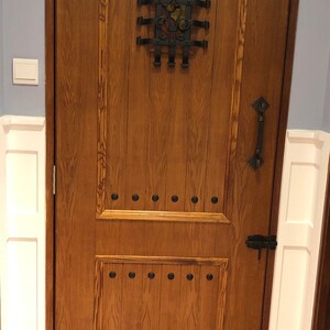 May include: A solid wood door with a dark brown stain. The door has a decorative metal grate with a fleur-de-lis design. The door has a black metal handle and a black metal latch. The door is surrounded by white trim.