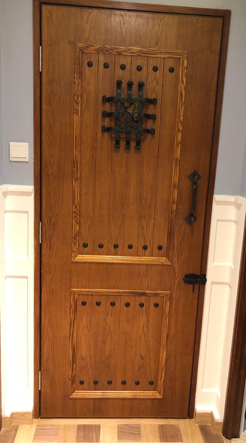 May include: A solid wood door with a dark brown stain. The door has a decorative metal grate with a fleur-de-lis design. The door has a black metal handle and a black metal latch. The door is surrounded by white trim.