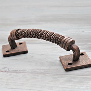 Könnte beinhalten: Ein bronzefarbener Schrankgriff mit einem geflochtenen Griff. Der Griff ist gebogen und an quadratischen Platten mit dekorativen Mustern befestigt. Die Hardware ist aus Metall und hat ein rustikales, antikes Aussehen.