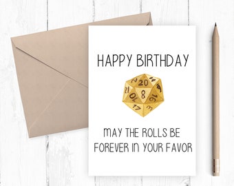 May include: A birthday card with the text "HAPPY BIRTHDAY" and "MAY THE ROLLS BE FOREVER IN YOUR FAVOR". The card features a gold-colored, twenty-sided die illustration. A tan envelope and a pencil are also visible.