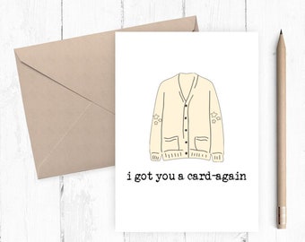 May include: A greeting card with a cream-colored cardigan illustration and the text "i got you a card-again". The card is accompanied by a tan envelope and a wooden pencil, all set against a white wooden background.