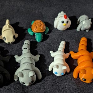 May include: A collection of 11 small, colorful, segmented animal figurines. The figurines are made of a soft, pliable material and are designed to be bendable. The animals include a turtle, a chicken, a cat, a dog, a fish, and a few others.