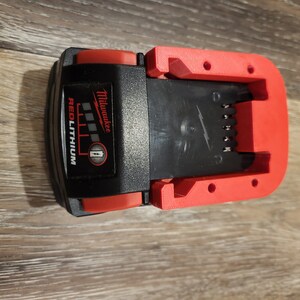 May include: A red and black Milwaukee REDLITHIUM battery charger with a black plastic base and red plastic sides. The charger has a slot for a battery and a power cord.