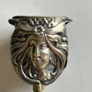 May include: A silver-toned pin with a detailed Art Nouveau design featuring a woman's face. The pin has a long, thin stem.
