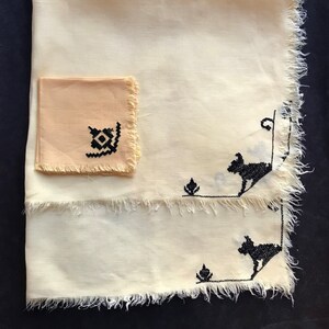 May include: A set of two white linen napkins with black embroidered designs. The larger napkin has two black dog silhouettes and the smaller napkin has a geometric design.