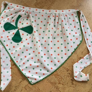May include: A white apron with green and orange polka dots and a green shamrock patch. The apron has a green and white trim around the edges.