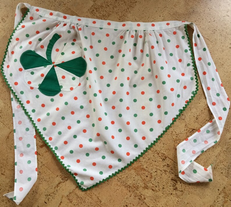May include: A white apron with green and orange polka dots and a green shamrock patch. The apron has a green and white trim around the edges.