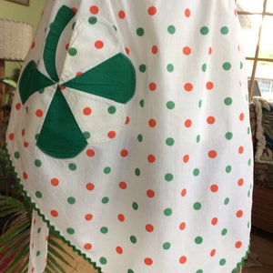 May include: A white apron with green and orange polka dots. The apron has a green and white patchwork pocket in the shape of a pinwheel.