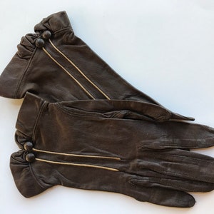 May include: A pair of brown leather gloves with gold stitching and black buttons. The gloves have a ruffled cuff.