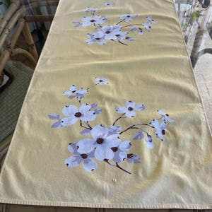 May include: Yellow rectangular table runner with a floral pattern. The design features white flowers with brown centers and light purple accents, arranged on branches. The runner is made of a soft, possibly linen, fabric.