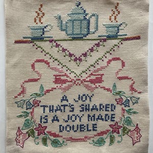 May include: Cross-stitch embroidery on beige fabric features a teapot, teacups, and floral accents. The text reads "A JOY THAT'S SHARED IS A JOY MADE DOUBLE." The design includes pink, blue, green, and brown thread, creating a vintage aesthetic.