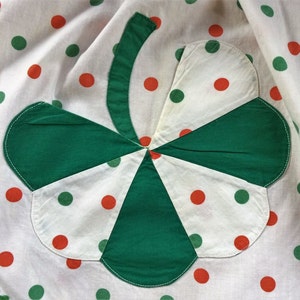 May include: A white fabric with green and orange polka dots features a large green shamrock sewn on top. The shamrock is made of four green fabric pieces sewn together.