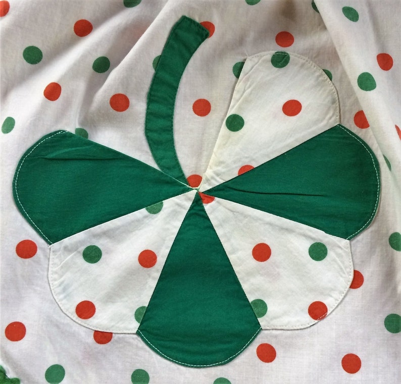 May include: A white fabric with green and orange polka dots features a large green shamrock sewn on top. The shamrock is made of four green fabric pieces sewn together.