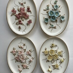 May include: Four oval wall plaques with raised floral designs. Each plaque features a different color scheme: pink, blue, pink roses, and yellow roses. The plaques have a white background with gold trim, and are a decorative home accent.