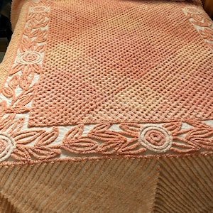 May include: A peach-colored bedspread with a raised floral pattern. The bedspread has a scalloped edge and a textured surface.