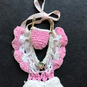 May include: A pink and white crocheted hanging with a small pink crocheted pouch and a wooden dowel. The hanging is decorated with a white ribbon.
