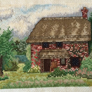 May include: A cross-stitch embroidery of a brick cottage with a thatched roof, surrounded by green trees and flowers. The cottage has a brown door and two windows.