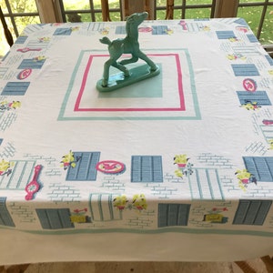 May include: White tablecloth with a blue, yellow, and pink repeating pattern of windows and flowers. A turquoise ceramic horse figurine is centered on the tablecloth.