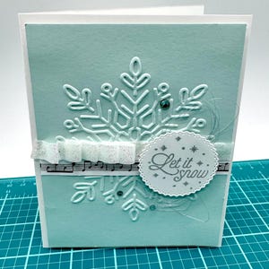 May include: A handmade greeting card with a light blue background and a large embossed snowflake. The card features a white ribbon, musical notes, and a circular tag that reads "Let it snow".